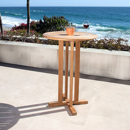 70682 Vogue teak 30 inch diameter Round Bar Table showing potted plant centerpiece and glass of wine on concrete terrace with shrubs and ocean view and blue sky background