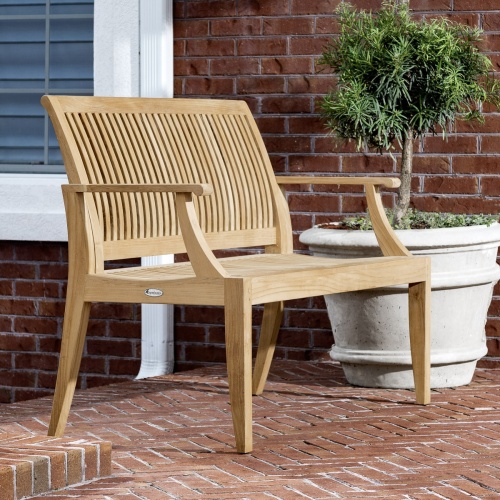 70778 Laguna six foot long teak bench angled on brick patio next to a potted tree in round white planter with brick wall and window on background