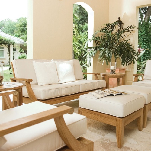 70877 Laguna 7 piece teak Love Seat Set on travertine tile patio open magazine on ottoman two pillar candles on side table potted palm tree with trees and house in  background