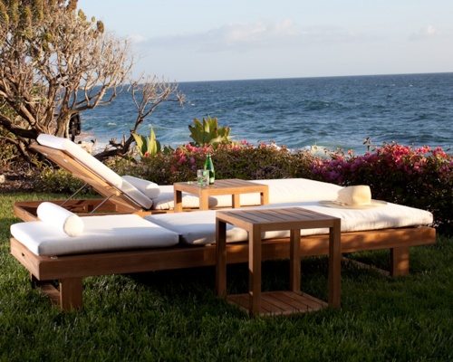 76770LM Horizon teak Lounger Cushion in Liso Marfil on 2 Horizon Loungers with 2 teak side tables with glass and bottled water on table angled on grass with ocean and blue sky