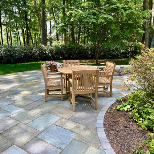 70483 Veranda teak 4 ft Round Dining Set of 4 Veranda Teak Armchairs and 4 foot round teak dining table on stone patio with plants and trees in background 