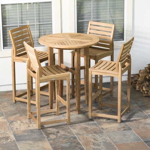 70532 Teak 5 piece Bar Table Set of 4 Somerset barstools and Laguna Table on tile patio against 2 window next to stack of chopped wood logs with house in background