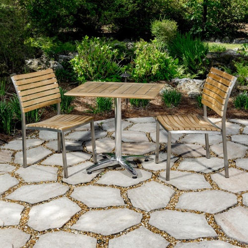 70665 Vogue Bistro Set of a teak and stainless steel rectangular table and 2 teak and stainless steel side chair on stone patio with landscape plants in background