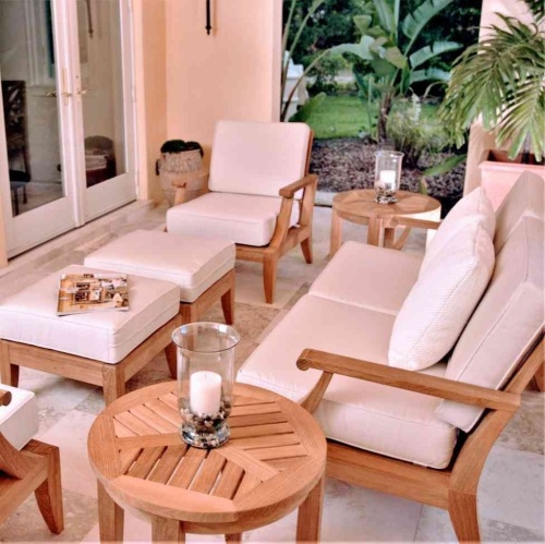 70877 Laguna 7 piece teak Love Seat Set on travertine tile patio open magazine on ottoman two pillar candles on side table potted palm tree facing doors to interior of house