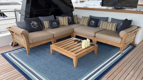 16767dp aman dais sectional set on a blue area rug on a boat deck featuring the Ottoman Fill teak base as a cocktail table with three pillar candles and two remotes boats in background