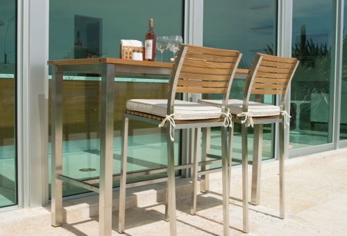 72000MTO Vogue teak and stainless steel bar set with seat cushions and 2 wine glasses and wine bottle and condiments and napkins on stone patio against glass wall angled side view 