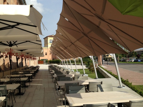 sps25100ffb spectra solo umbrella only on a restaurant outdoor dining area next to paved sidewalk with grass trees buildings and parking lot in background 