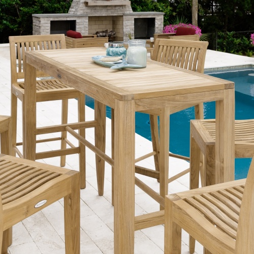 15821 Laguna 5 foot Teak Bar Table and 4 barstools on patio stones showing candle fish platter and jar on table next to pool with fireplace and flowering plant in background