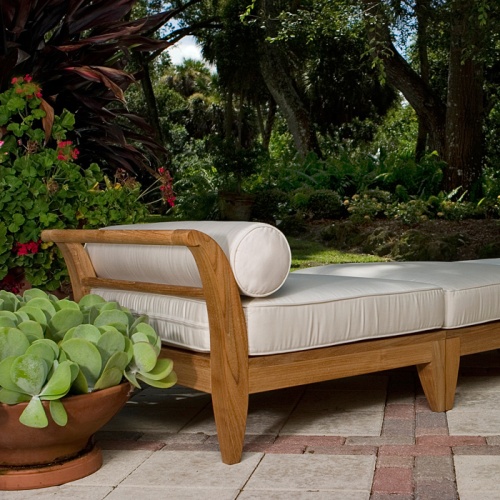 16766 aman dais teak end base and ottoman set angle view on a outside paver patio with a bolster and cushions with a container of green succulents in background of lush green shrubs
