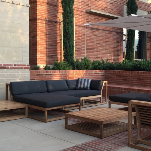 16800DP Maya Teak Sectional Sofa set with black cushions and throw pillows two cocktail tables on concrete patio with brick planter with cypress trees and plants with brick wall background