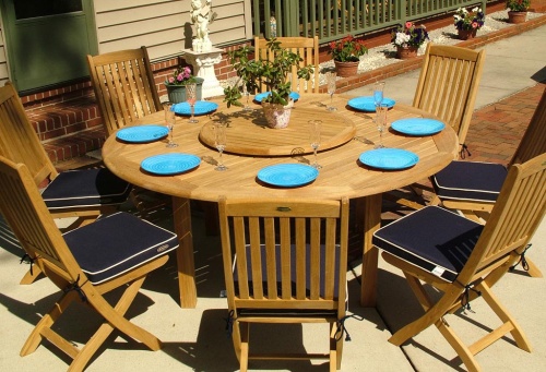 70018 Buckingham Barbuda 9 piece teak round dining set with 8 blue plates champagne glasses a potted plant on optional lazy susan on concrete patio surrounded by potted plants 