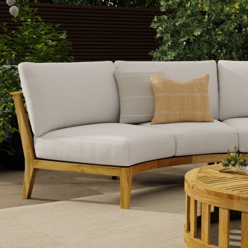 70881 Kafelonia Teak 7 pc Lounge Set in partial view of Sofa Sectional Bench with 2 throw pillows on an outdoor patio with coffee table in front of landscaping shrubs in background