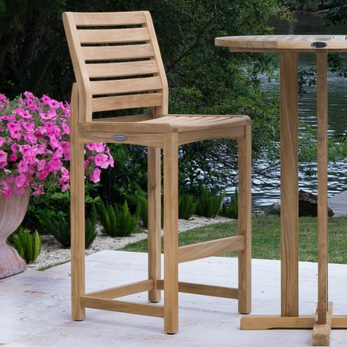 70514 Somerset teak barstool side angled with round bar table on concrete patio beside potted flowering plant with grass landscape plants shrubs and lake in background