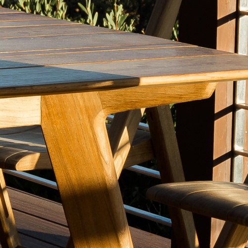 70524 Surf Horizon teak Dining Set closeup view of side of table top and table leg on wood deck with balcony and shrubs in background