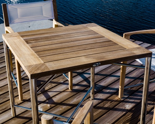 70459 Odyssey folding teak and stainless steel dining table and chair on dock with water in background