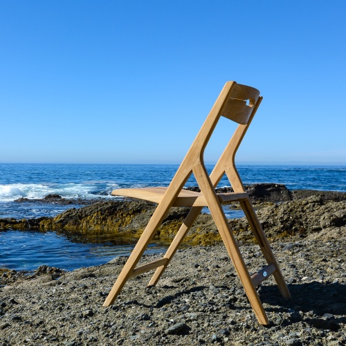 70616 Surf Teak Dining Chair on sandy beach facing the ocean with large boulders and blue sky in background