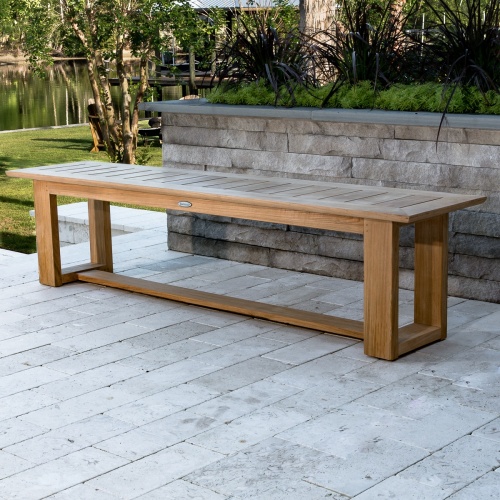 13909RF Refurbished Horizon teak 6 foot long Backless Bench angled view on paver patio next to paver planter with landscape plants and green grass and trees and waterway in in background 