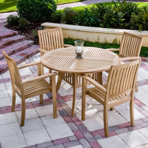  12196 sussex teak stacking armchair with round teak dining table on stone patio with trees and shrubs in background