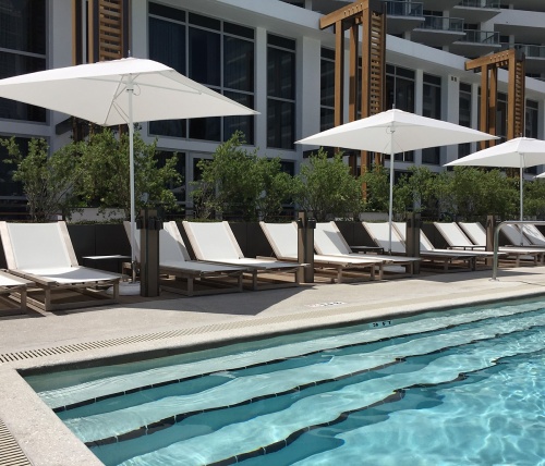 16771dp Maya teak Sling Lounger in white textilene mesh fabric and three market umbrellas on a concrete pool deck lined with trees facing the pool with eden roc resort in the background