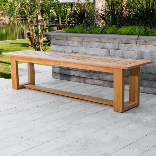 70497 Horizon teak Picnic backless bench on outdoor patio against paver planters with plants  grass area trees shrubs and lake in background