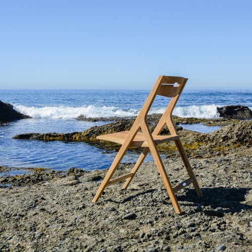 70672 Surf teak folding chair on sandy beach with large rocks in background facing ocean and blue sky