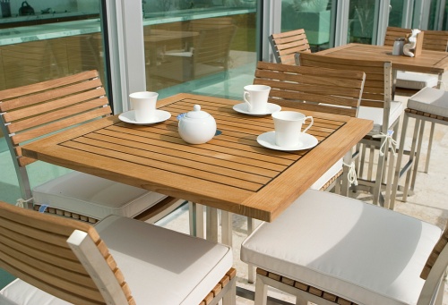 25313 Vogue teak and stainless steel Bar Table and 4 chairs with optional cushions on concrete patio teapot and 3 cups napkin holder salt pepper with glass sliding doors in background