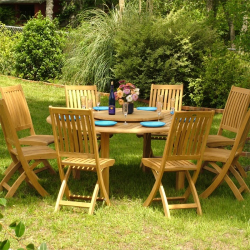 70018 Buckingham Barbuda 9 piece teak round dining set with 8 blue plates champagne glasses a flower vase on optional Lazy Susan on grass with plants and trees in background