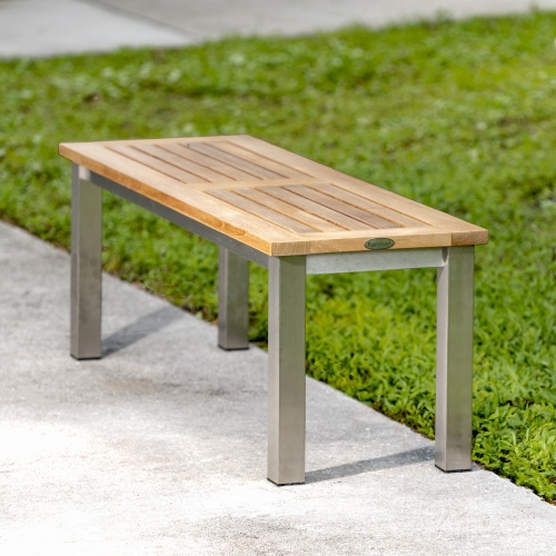 23940  Vogue Teak and Stainless Steel 4 ft Backless Bench angled end view on concrete walkway with grass in background
