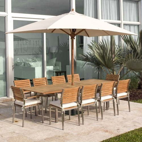 70176 Vogue stainless steel and teak 11 piece rectangular dining set with optional seat cushions and optional open market umbrella on stone patio with windows in background