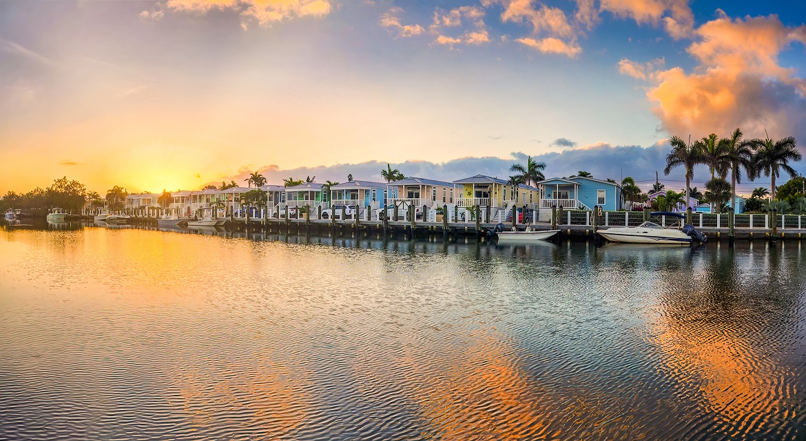 An image of a tropical sunrise in Key West with residential homes with palm trees and a dock showing seven docked boats overlooking the ocean.
