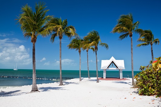 An image of a white gazebo with palm trees and sea grape shrubs on the beach with the ocean and blue sky background in Key Largo, Florida.