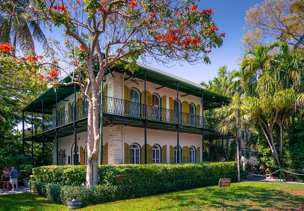 An image of Ernest Hemingway’s home in Key West in lime-colored Spanish Colonial style with covered porch with wooden shutters and a tropical garden landscape.