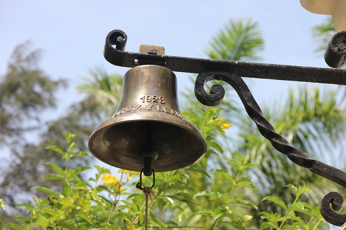  An image of the metallic bell at Ernest Hemmingway’s House with trees and blue sky in the background.