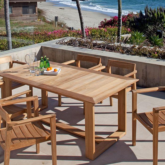 alt="70299 Horizon teak 7 piece Dining Set with 3 sparkling water bottles a water pitcher and plate of oranges on concrete terrace with landscape plants in concrete planter"