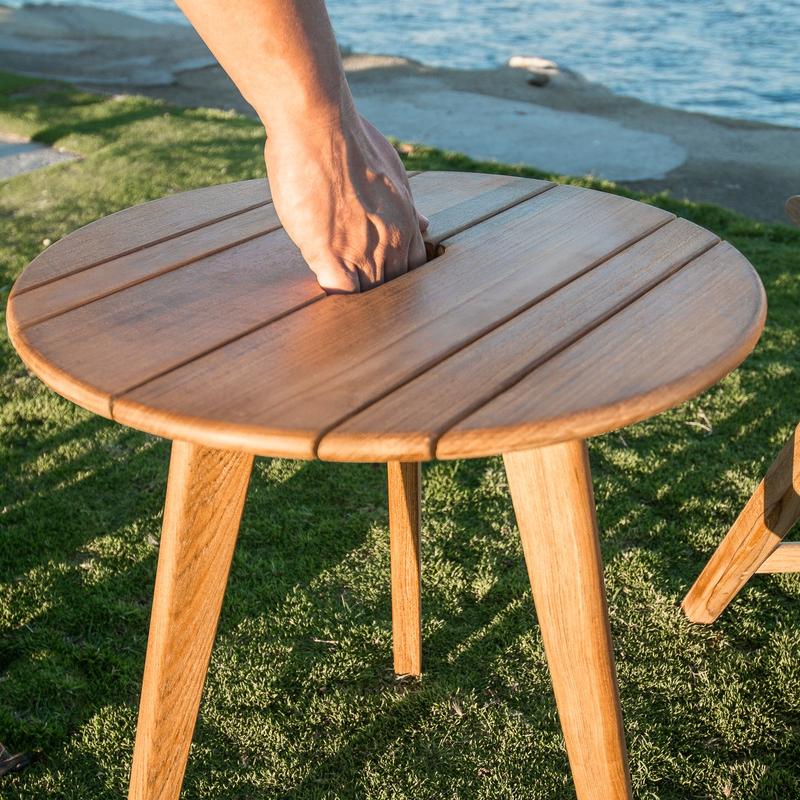 alt="14916 surf teak side table showing hand in carrying handle on grass with sand and water in background"