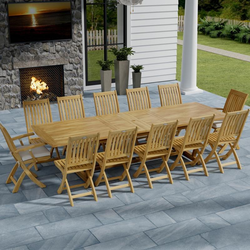 alt="70059 Grand Barbuda 13 pc Teak Dining Set with optional seat cushions in angled aerial view on patio in front of an outdoor fireplace and potted plants with house and lawn in background"