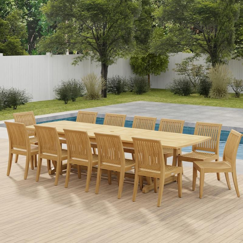alt="70124 Grand Laguna 13 pc Teak Side Chair Set of Grand Veranda Dining Table and 12 Laguna Side Chairs in angled side view on a patio in front of a pool with plants and fence in back"