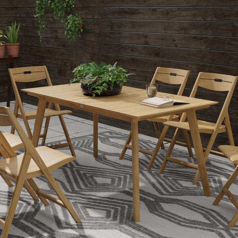 alt="15917 Surf Teak Dining Table with 6 Surf Chairs on area rug showing a plant and open book with glasses on table top and a wood fence with hanging plants in background"
