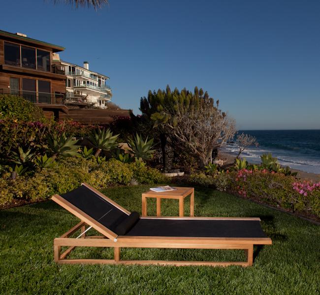 alt="16771 Maya Chaise Lounger in tobacco Textilene mesh fabric teak side table with magazine on a green lawn overlooking the ocean with flowering plants and two houses in background "