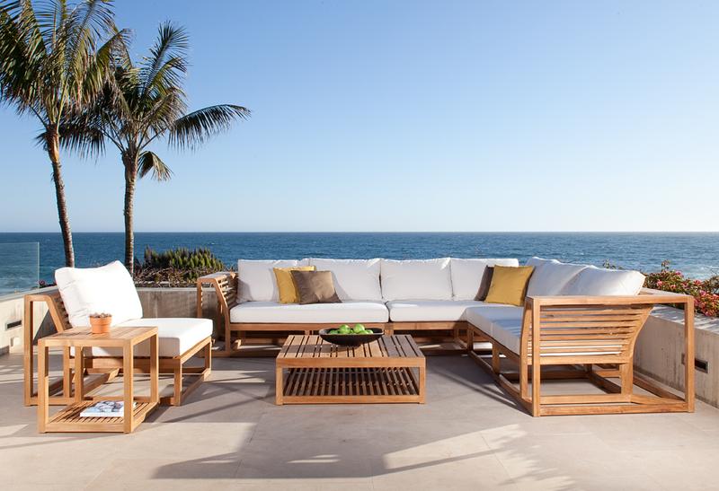alt="11800RFDP Maya teak lounge set with bowl of apples a potted plant and magazine on table shelf on concrete patio surrounded by two palm trees and plants with ocean and blue sky background"
