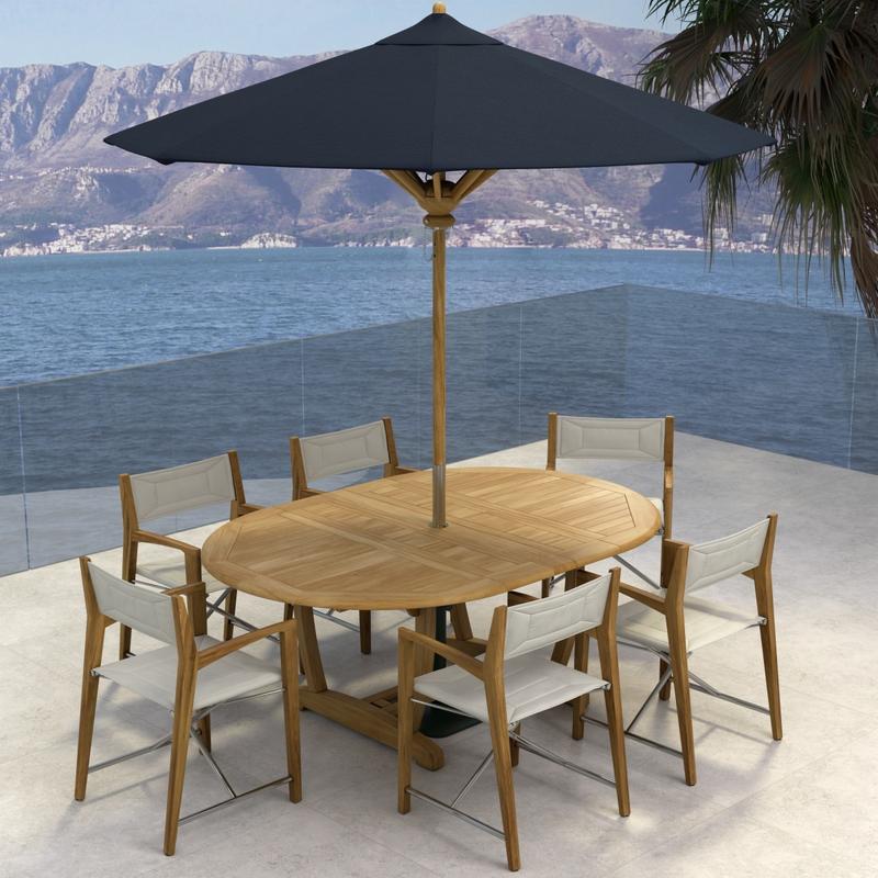 alt="70366 Oval Odyssey Chair 7 pc Dining Set angled aerial view with opened optional black umbrella on outdoor terrace next to palm tree with the ocean and mountain background "