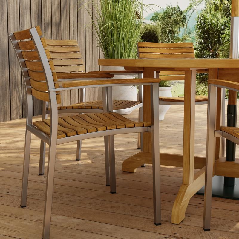 alt="70349 Barbuda Vogue Teak 7 pc Dining Set showing closeup view of Vogue Teak and Stainless Steel  Armchairs in side view on paver patio with wood privacy wall and trees in background "