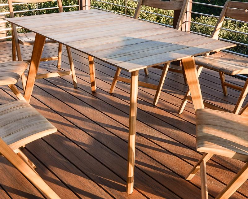 alt=" 70552 Vogue Surf teak 5 foot rectangular dining table closeup view of dining table on wood deck with plants in background"