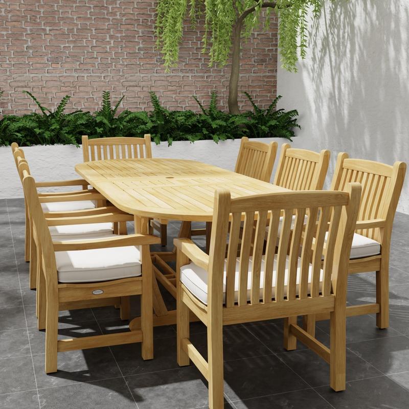 alt="70178 Montserrat Veranda Teak 9 pc Dining Set of Montserrat Dining Table and 8 Veranda Side Chairs with optional seat cushions in end view on patio with planter of ferns and tree in the background"
