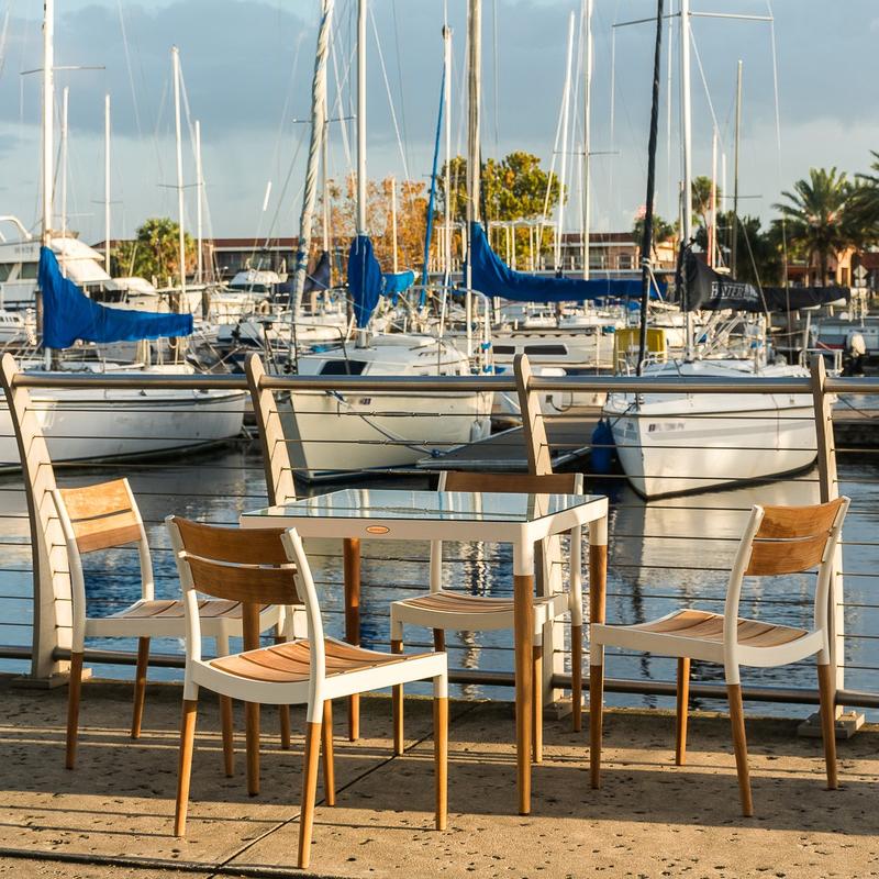 alt="70449 Bloom Glass Top Dining Set of Bloom teak and powder coated aluminum 32 inch square dining table on boat dock and marina with boats and palm trees and blue sky in background"