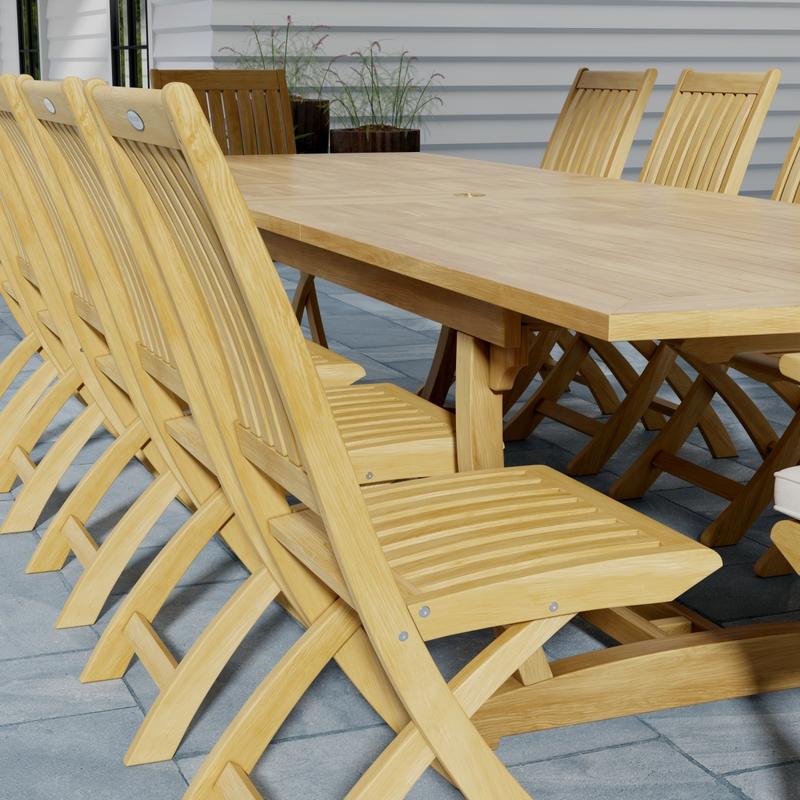 alt="70059 Grand Barbuda 13 pc Teak Dining Set showing closeup end view of table and side chair on patio with plants in the background"