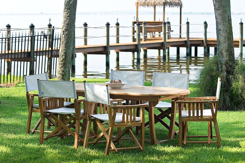 alt="70079 Director Chair 7 piece Oval Dining Set on grassy area with white bowl of pears on table 2 trees metal fence boat dock and lake in background"