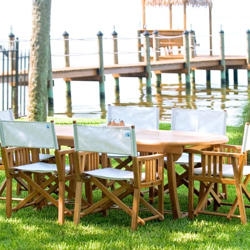 alt="70015 Montserrat Director 9 piece teak Dining Set with bowl of pears on grass lawn with boat dock and lake in background"