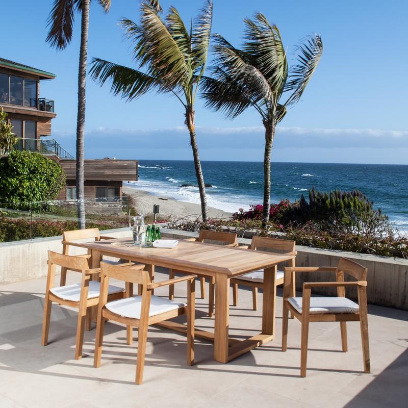 alt="70299 Horizon teak 7 piece Dining Set with bottles sparkling water a water pitcher on table showing optional seat cushions on terrace surrounded by plants with ocean and blue sky in back"