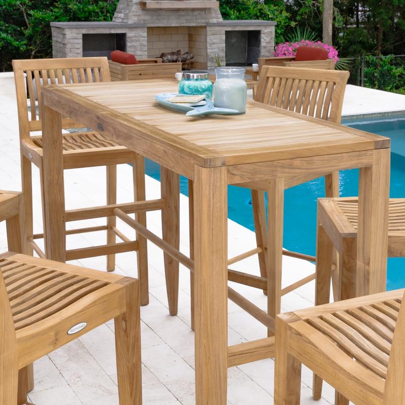 alt="70500 Laguna 5 piece Teak High Bar Set on patio pavers showing 2 blue candles and fish plate on table by pool with fireplace 2 chairs potted flowering plant and shrubs in background"